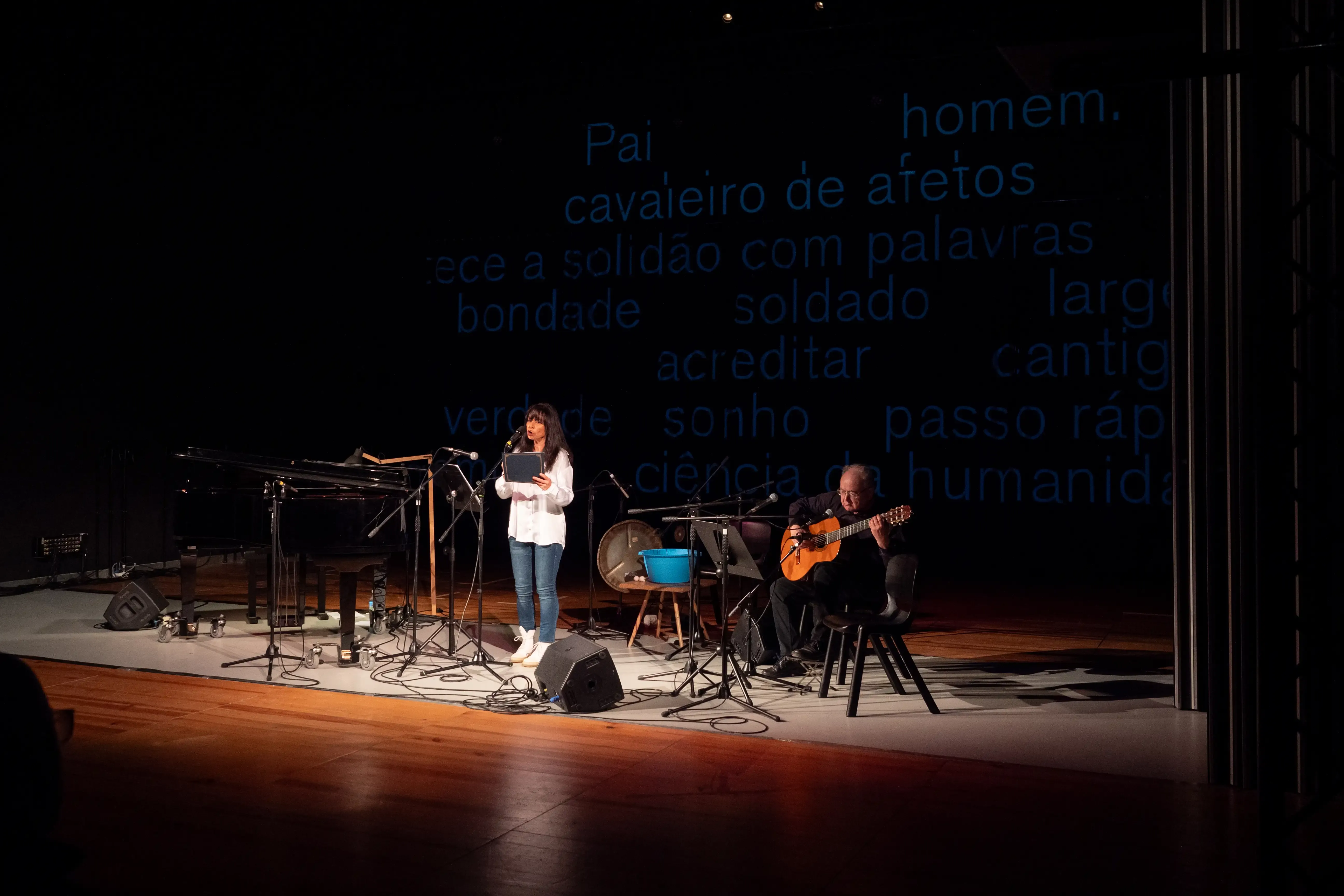 Uma mulher está a performar num palco. No lado direito da imagem um homem acompanha-a na guitarra clássica. Ao fundo estão projetadas palavras dispersas azuis.